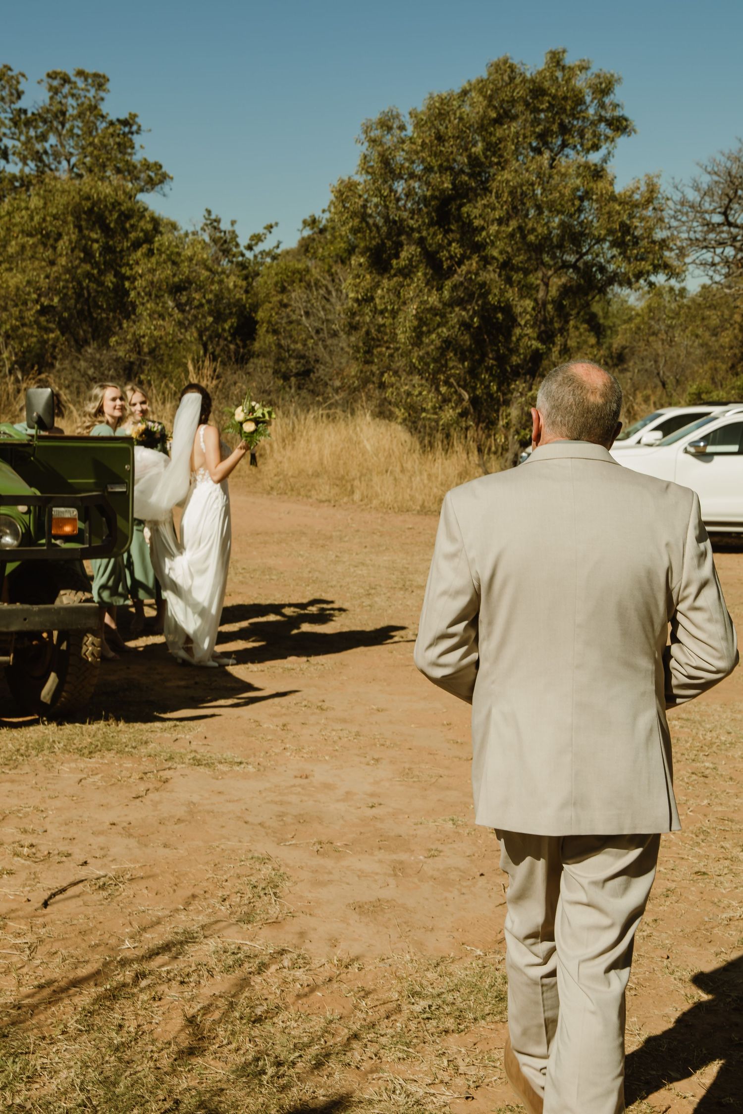 Wedding guests in formal attire walking along a dusty outdoor path with safari vehicles in background.