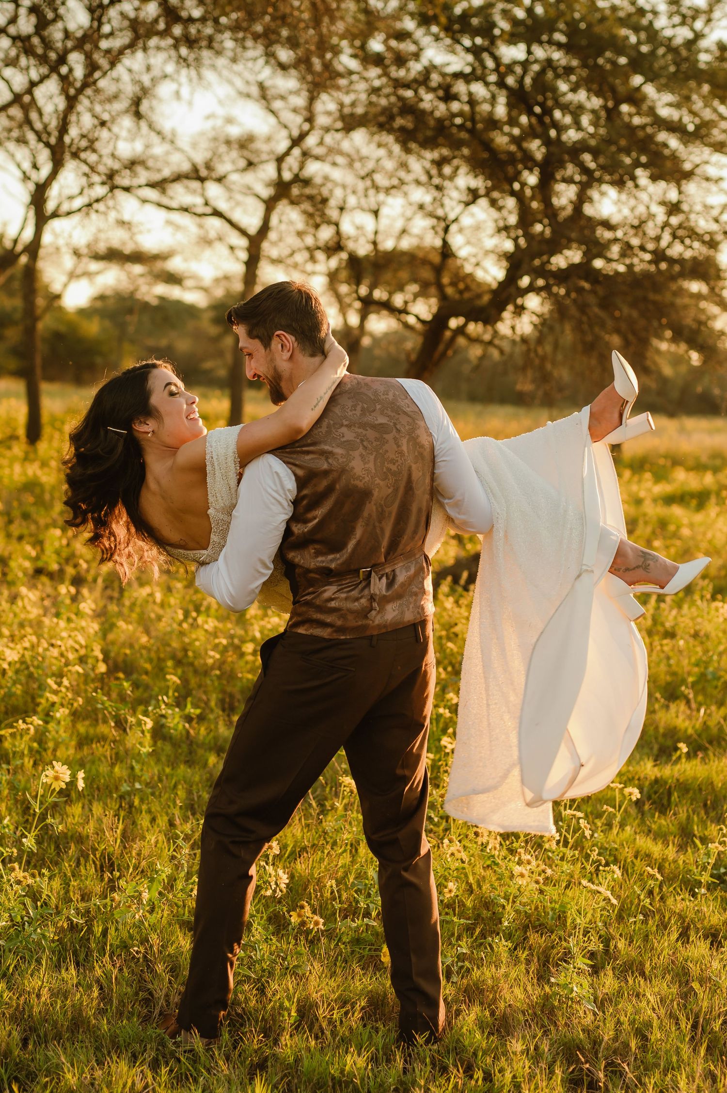 A romantic couple dances in a golden field at sunset with trees in the background.