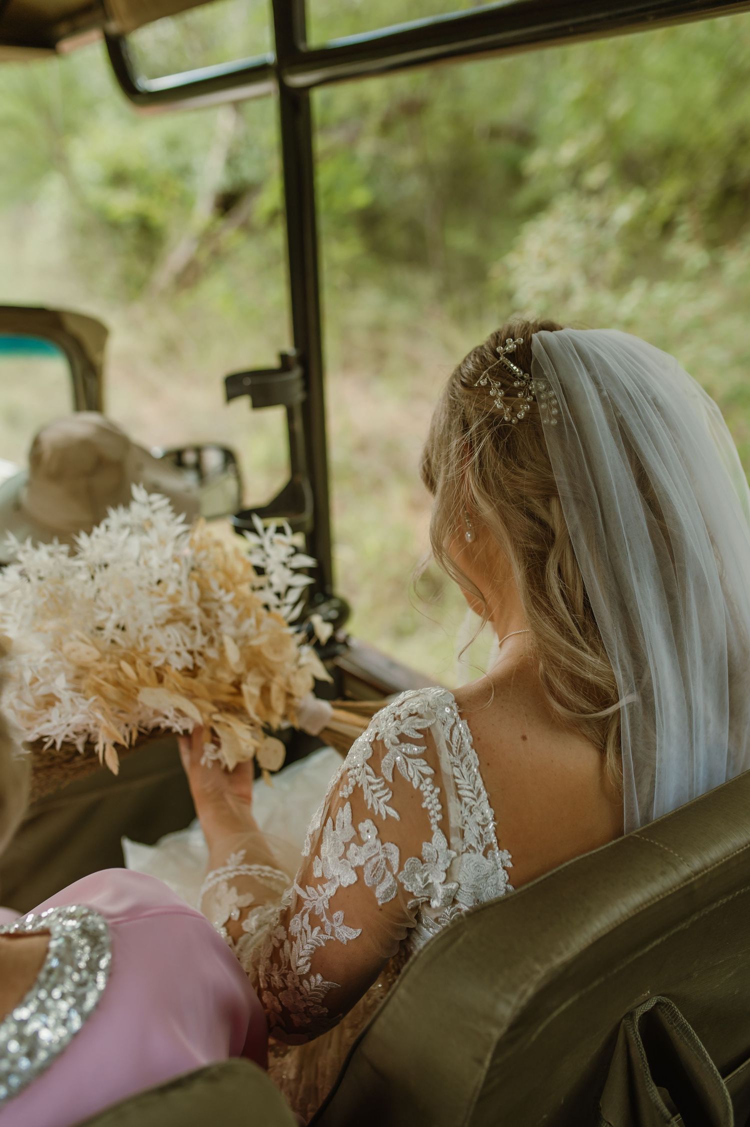 A bride in a lace dress with a veil holds a dried floral bouquet while sitting in a vintage car.