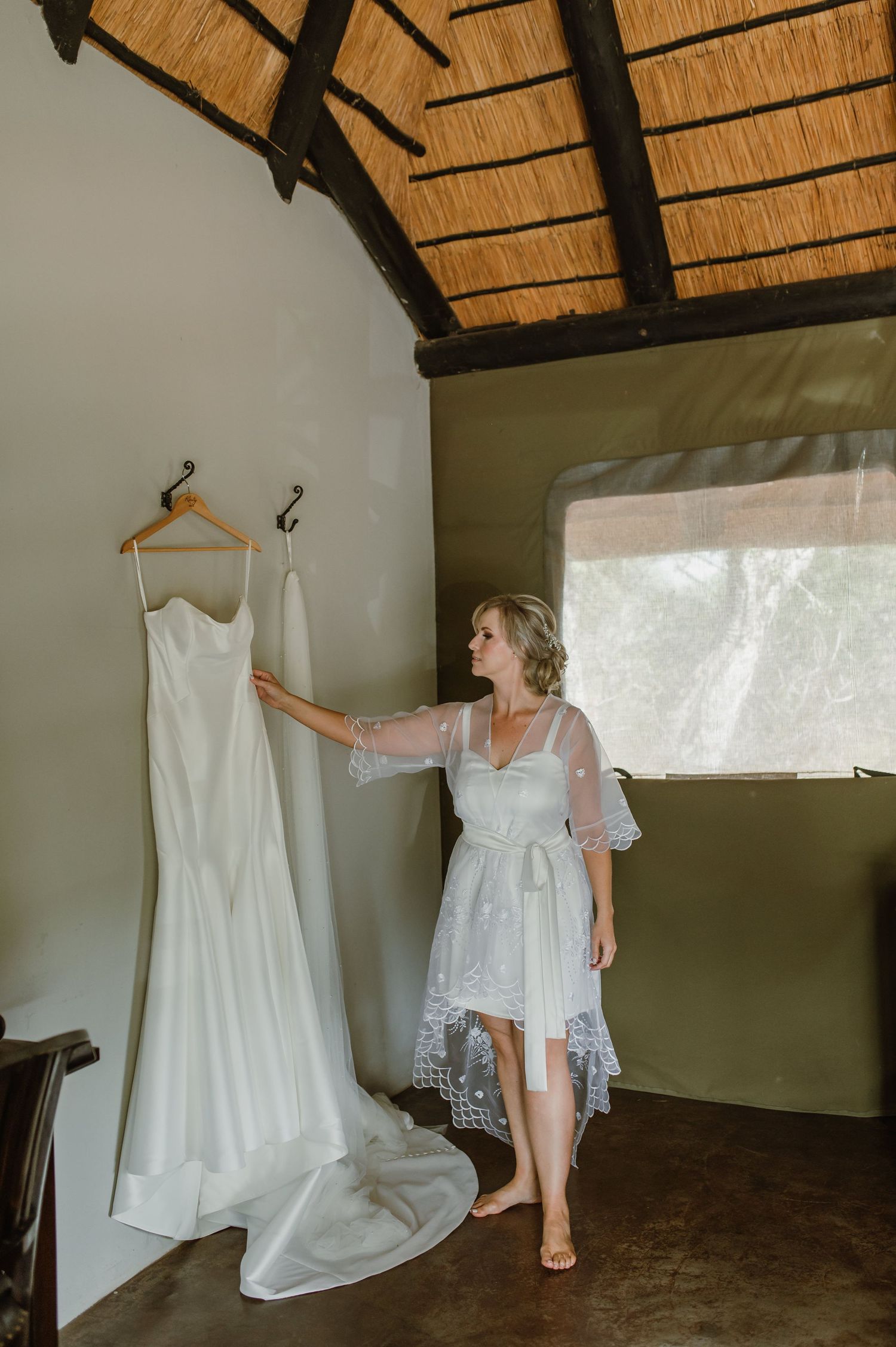 Wedding dress displayed in rustic room with thatched roof and natural lighting.