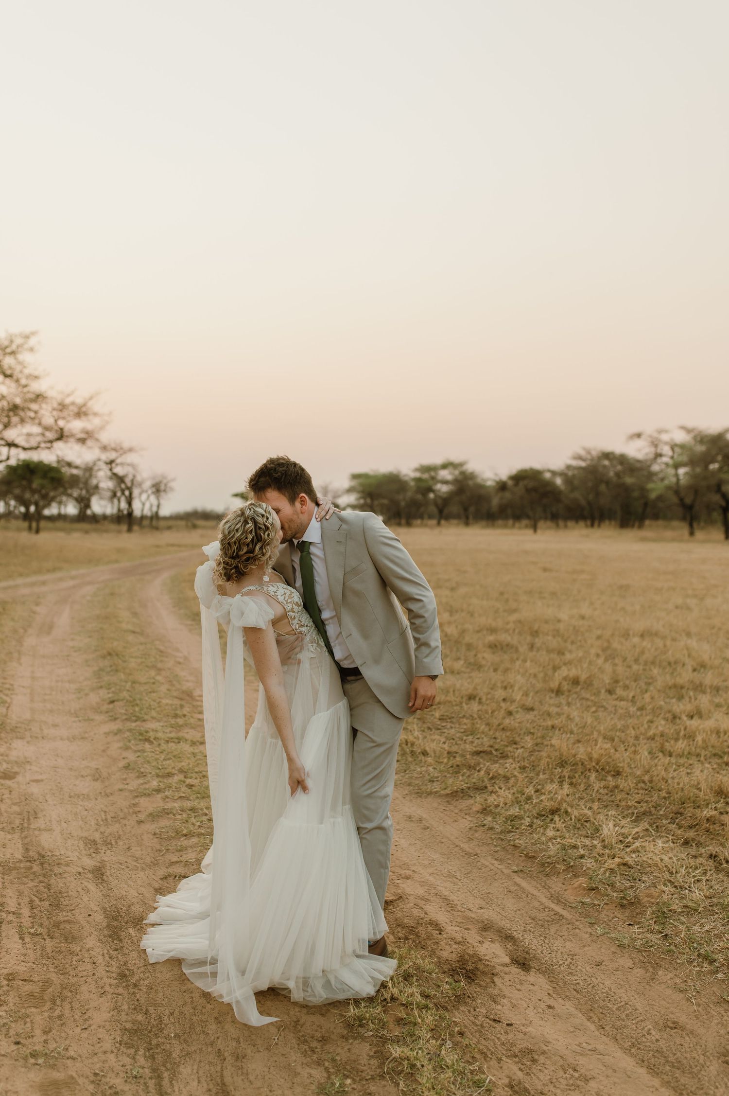 Bride in lace dress and groom in grey suit share playful moments on a rustic dirt road at sunset.