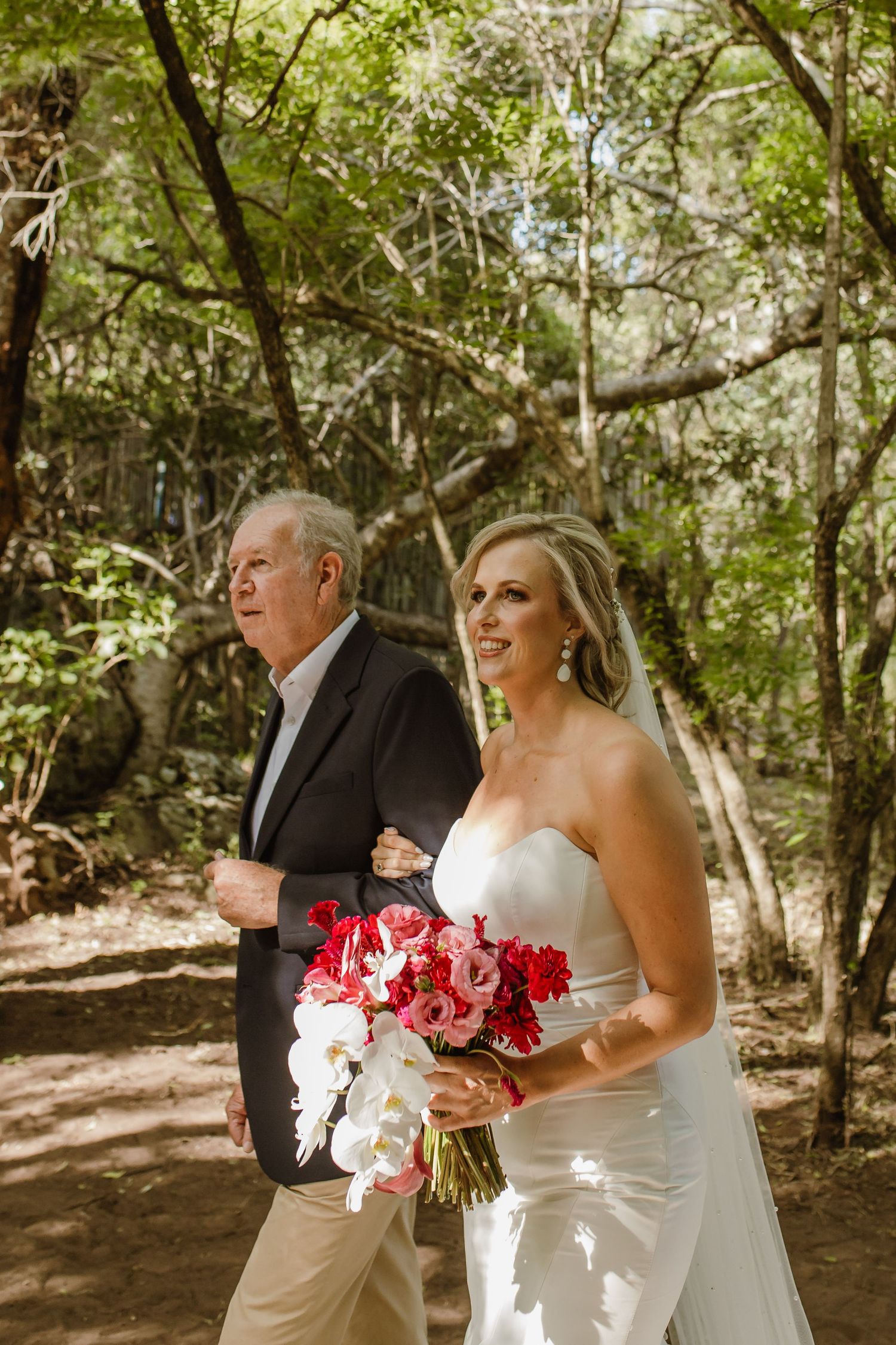 A bride in white holds a vibrant red and white bouquet while walking down a wooded path.