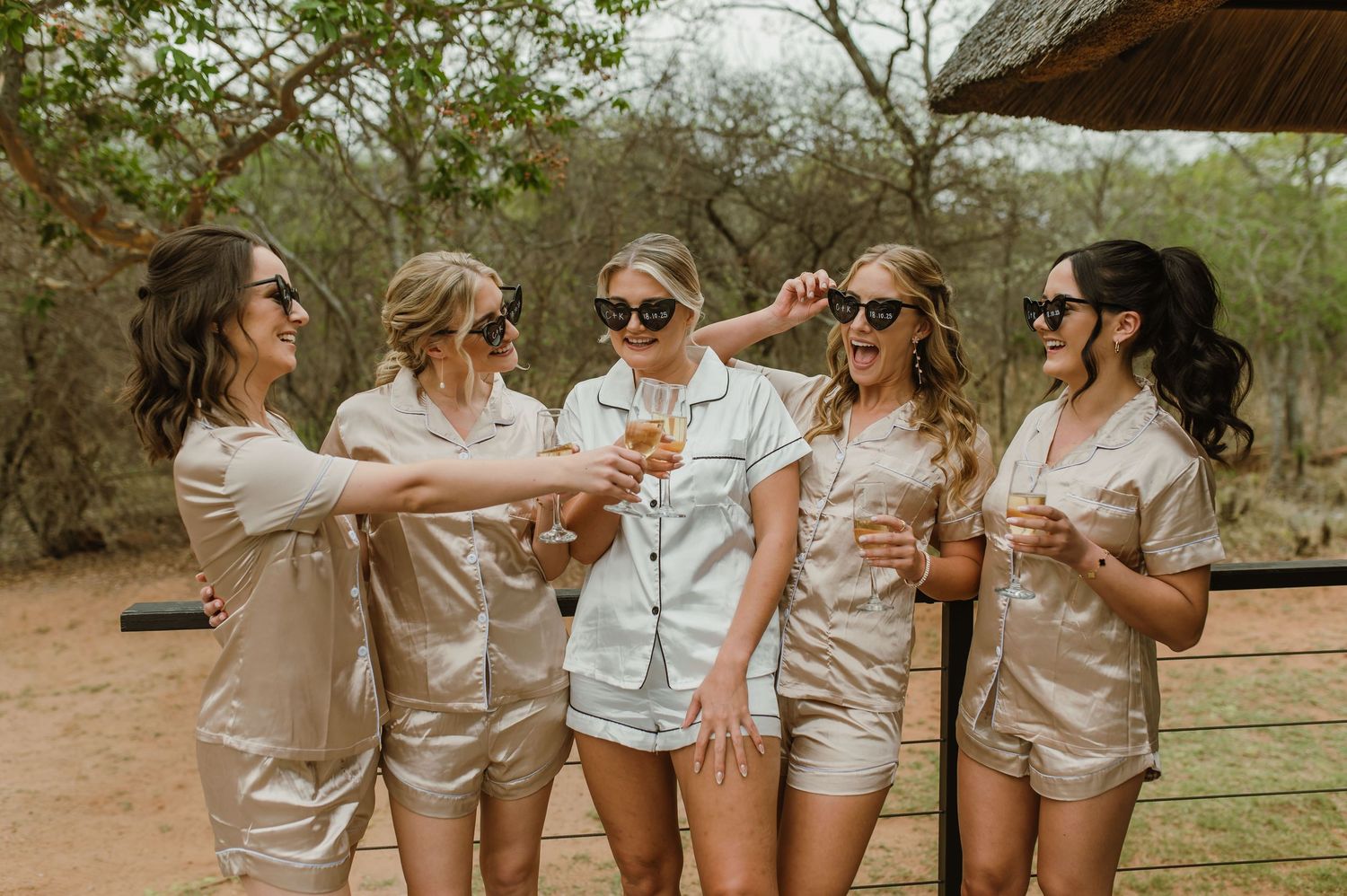 Friends in matching silk pajamas toasting with champagne glasses on outdoor deck.