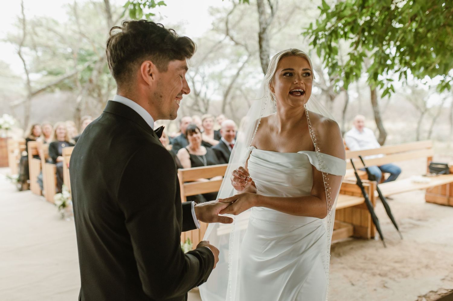 Couple shares a romantic moment during their outdoor wedding ceremony under green trees.