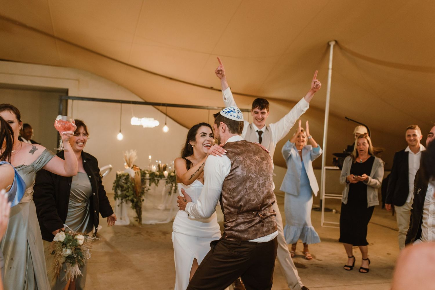 Guests dance and celebrate at a wedding reception under a tent with string lights and floral decorations.