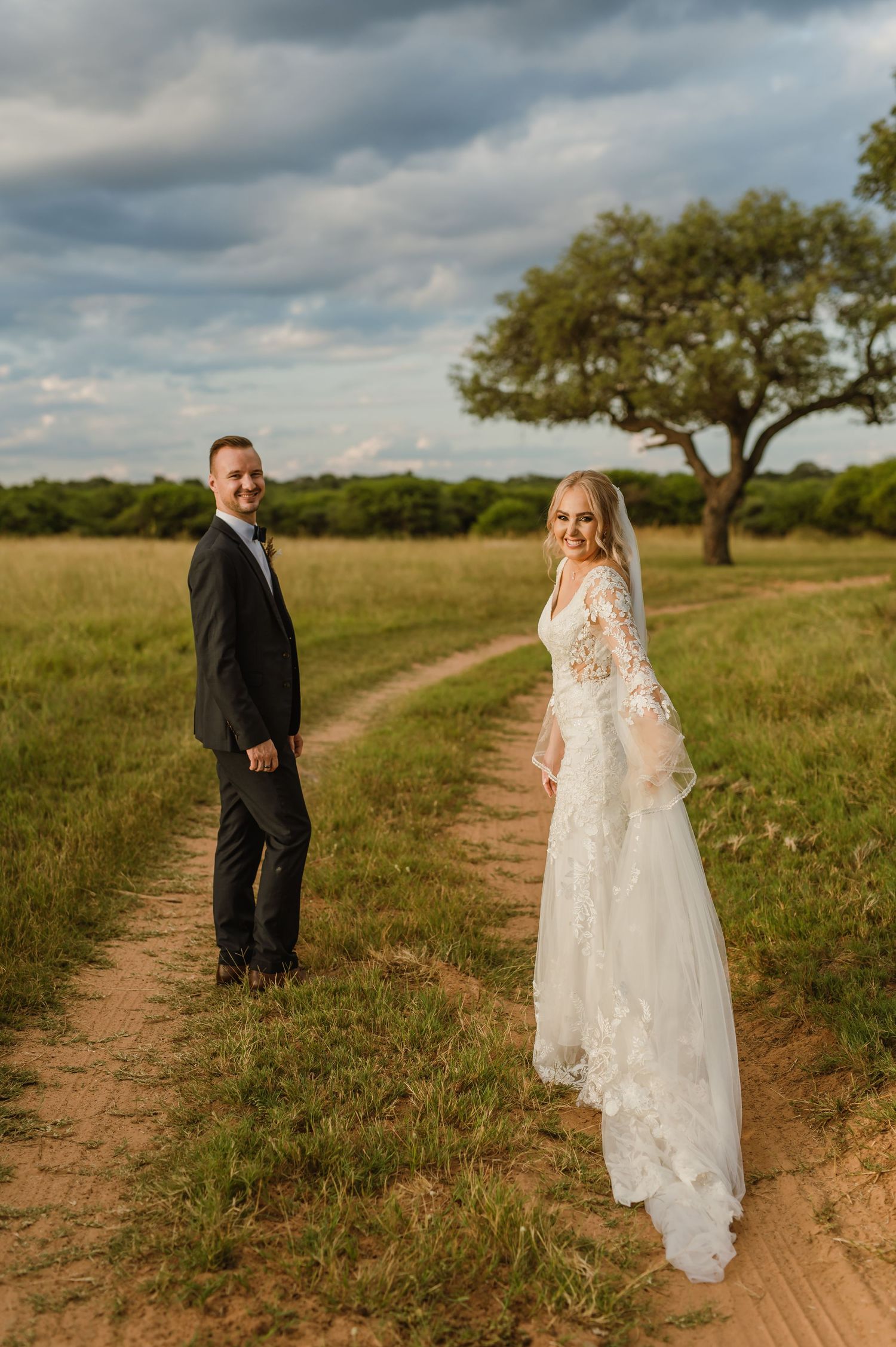 Couple in wedding attire walking along a dirt path through a rustic ranch landscape.