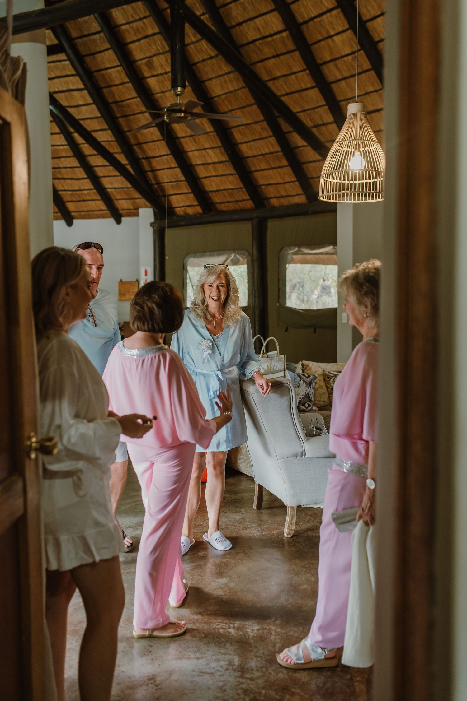Wedding party in pink and white robes socializing in a thatched-roof venue.