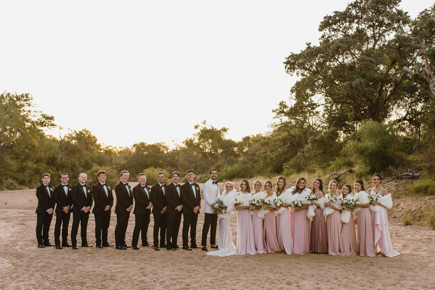 Large wedding party in formal attire poses together on sandy beach at sunset.