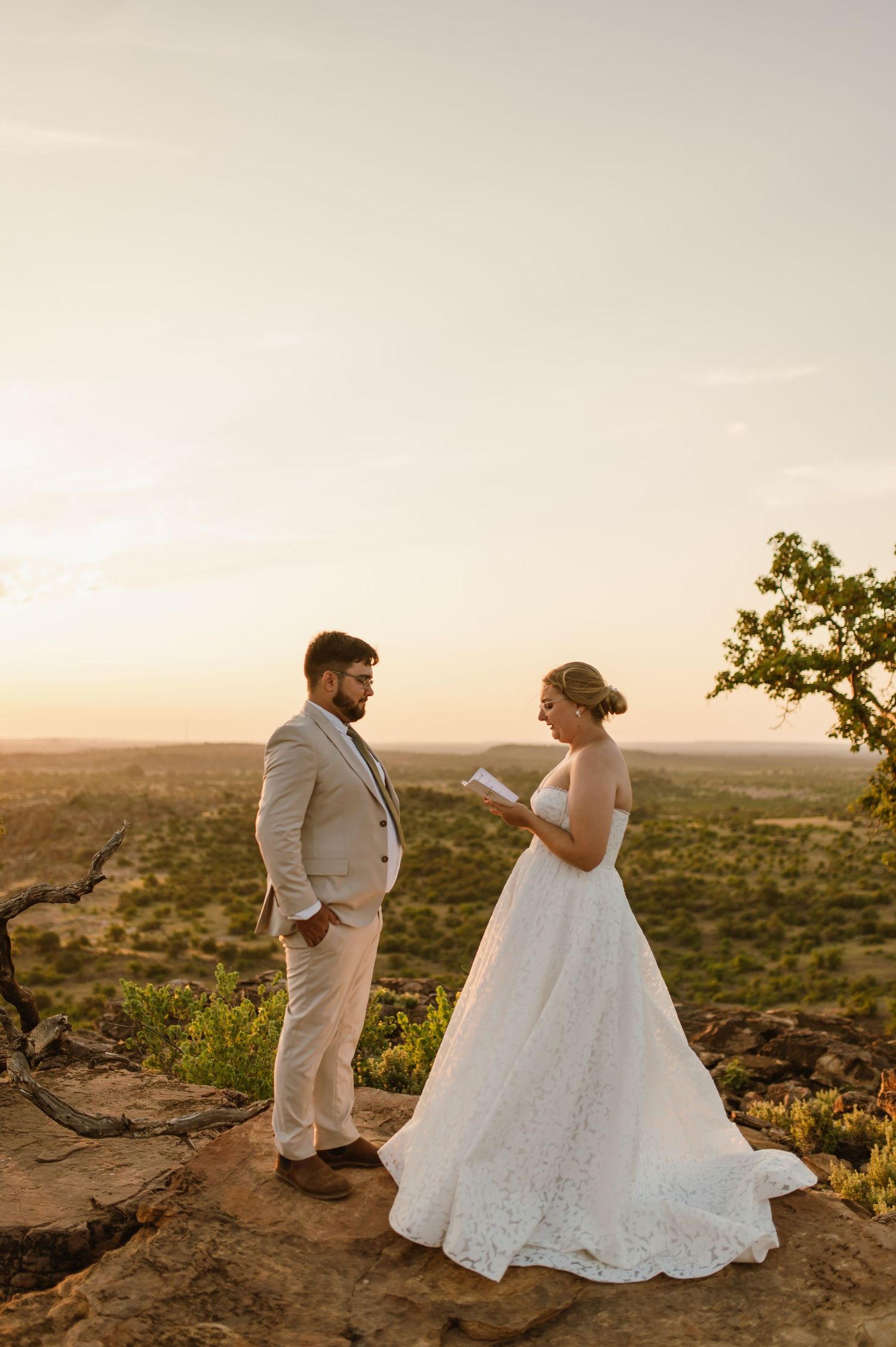 A couple in wedding attire stands on a scenic cliff edge overlooking a vast desert landscape at sunset.