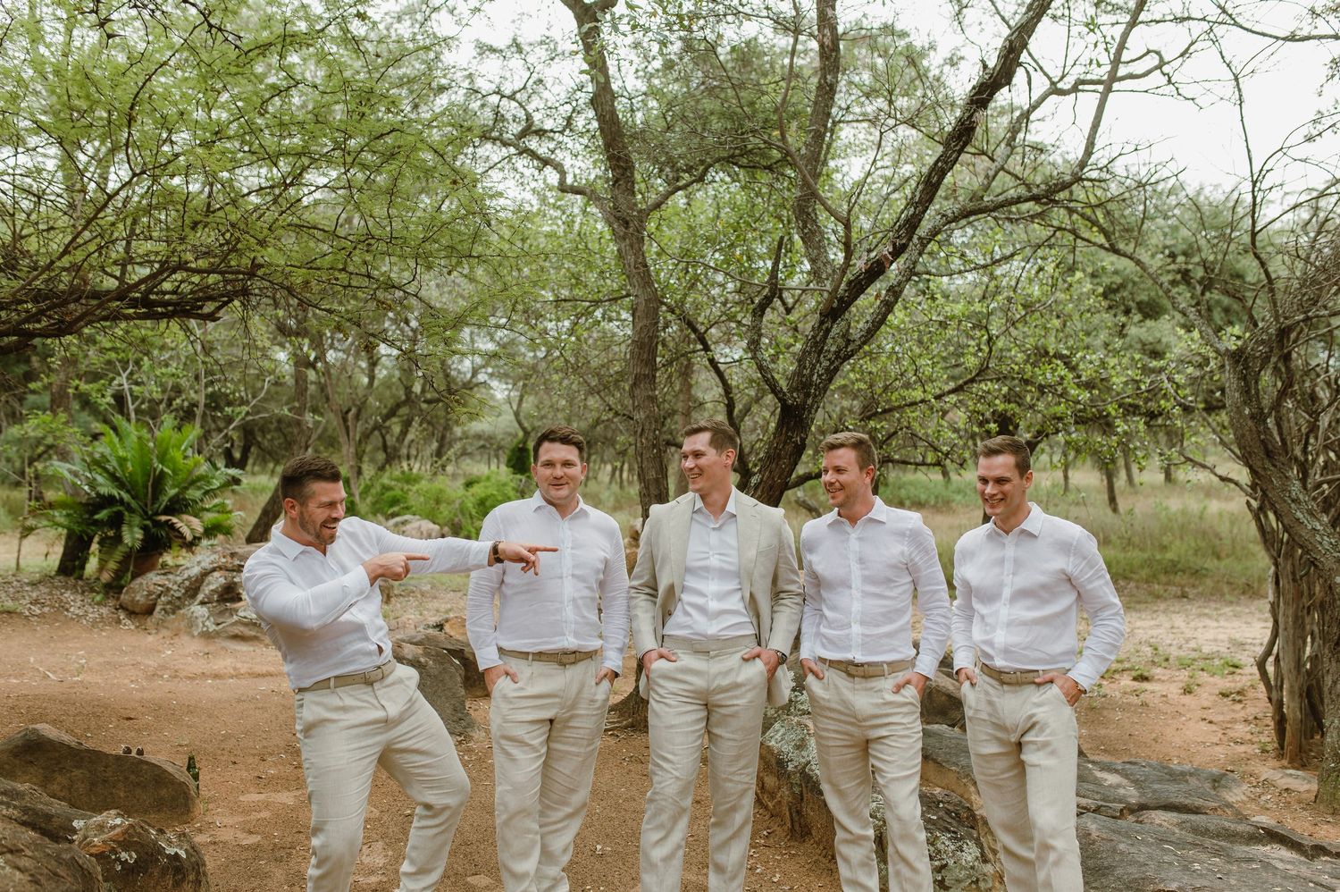A group wearing matching white outfits poses together in a natural outdoor setting with trees and greenery.