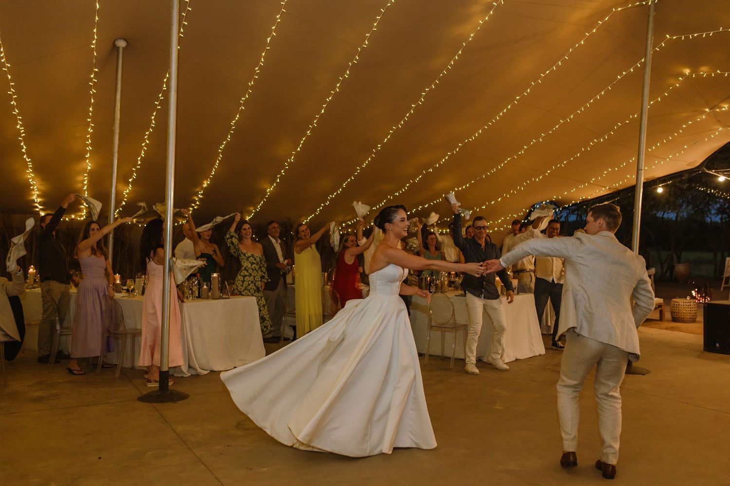Wedding celebration with guests dancing under a tent decorated with warm string lights.