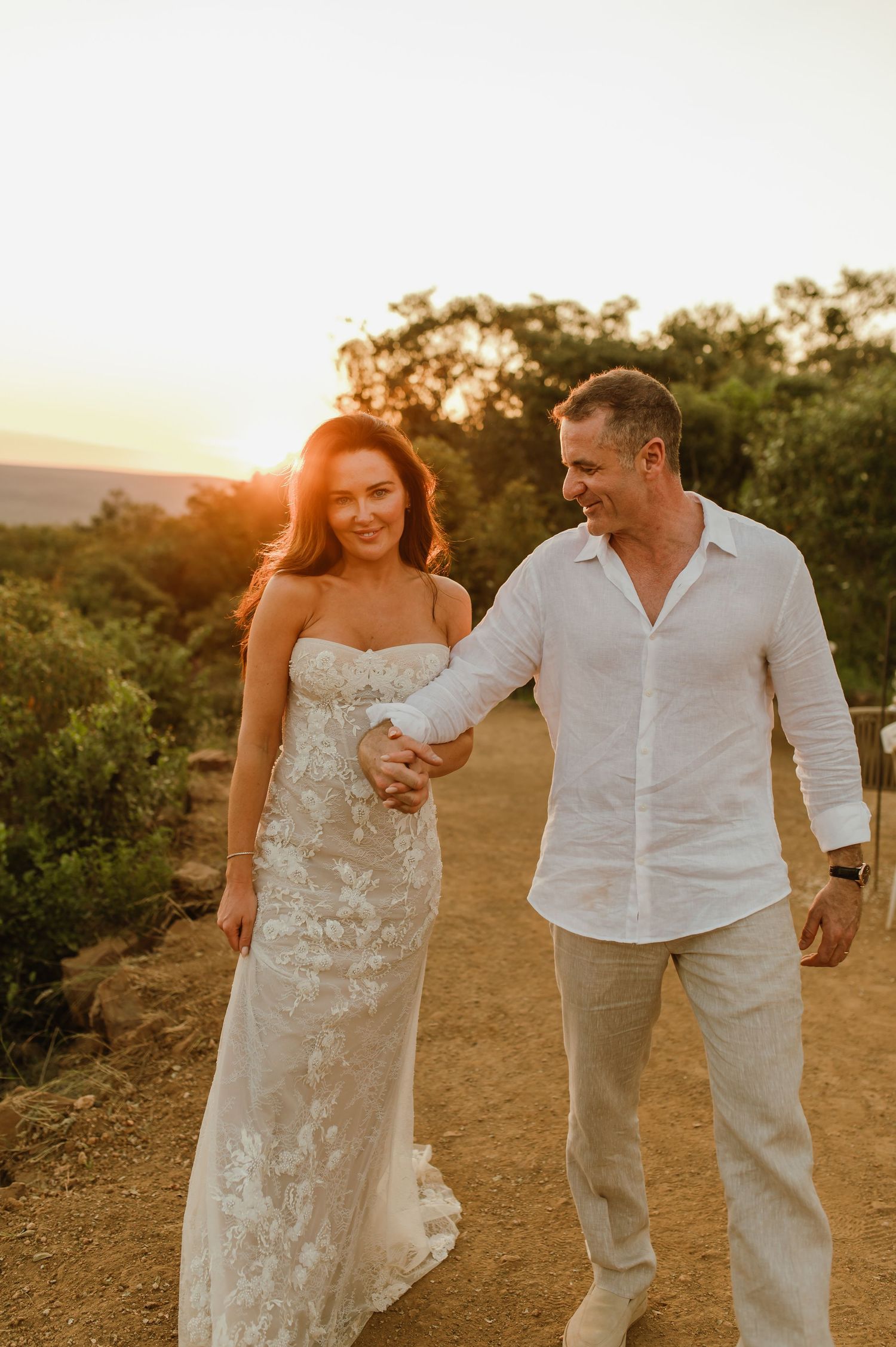 Newlyweds walk hand in hand on dirt path at sunset, bride in lace gown smiling.