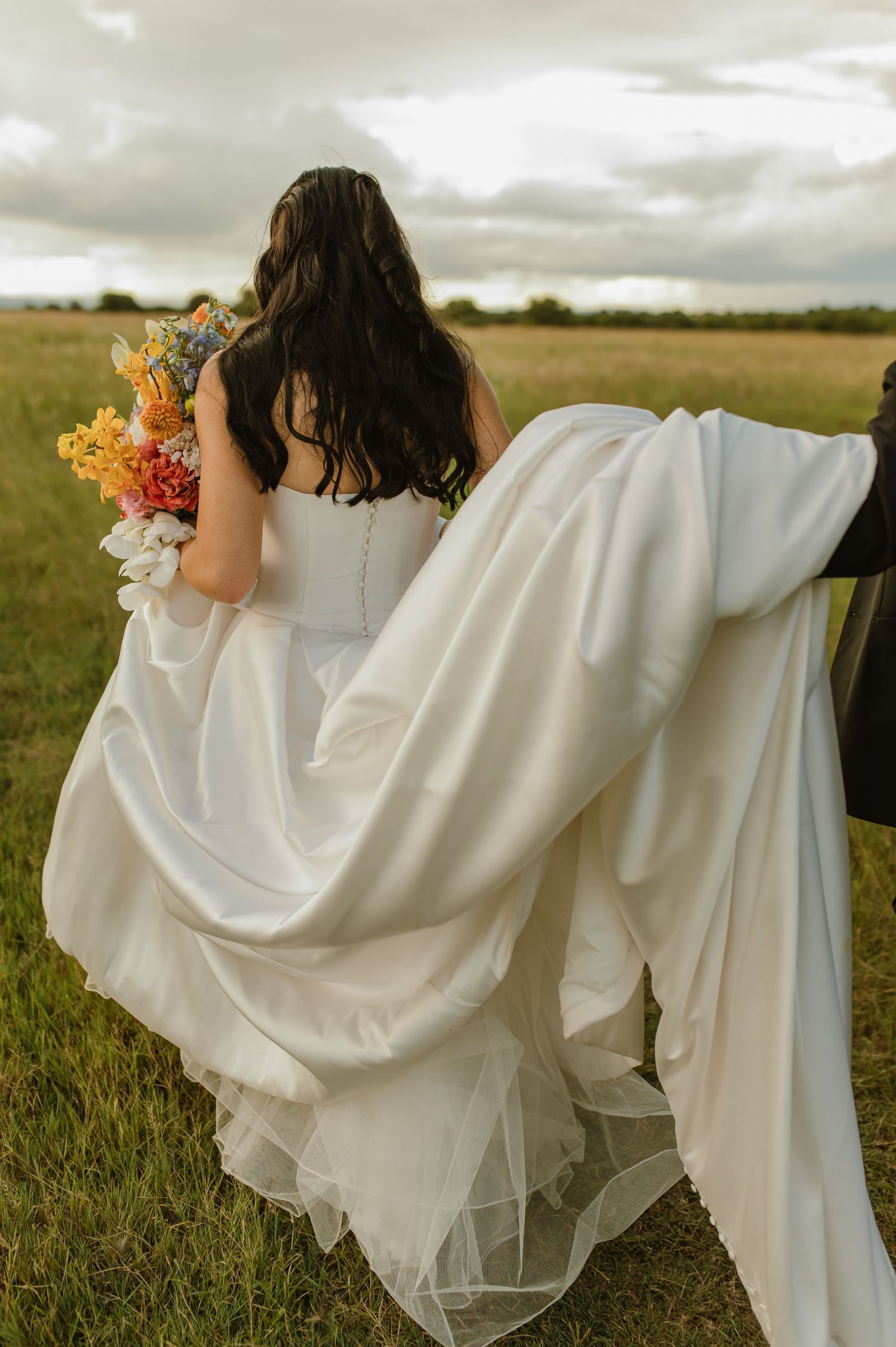 Bride in white satin gown holding colorful bouquet walks through green field under cloudy sky.
