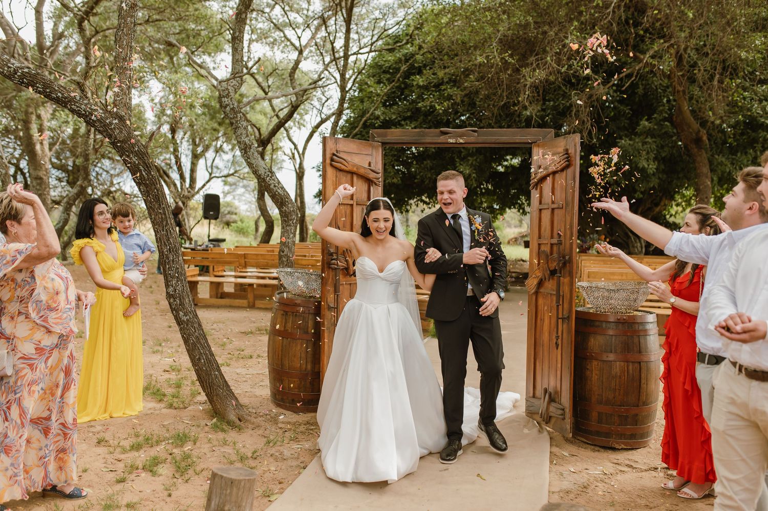Bride and groom exit ceremony through rustic wooden doors as guests celebrate around them.
