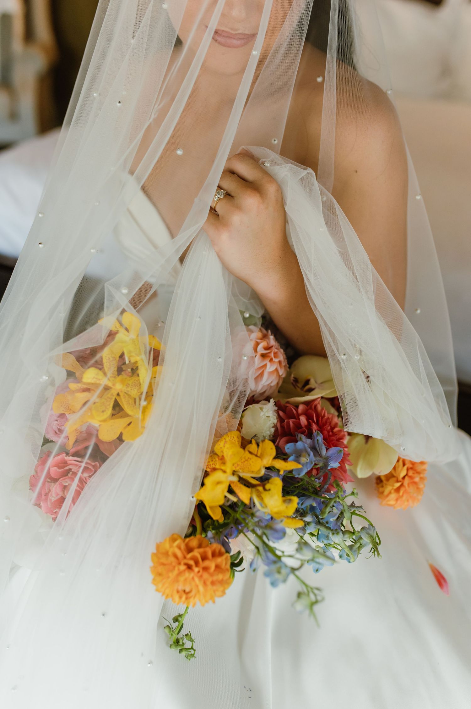 Bride in pearl-embellished veil holding colorful wildflower bouquet with yellow orange and blue blooms.