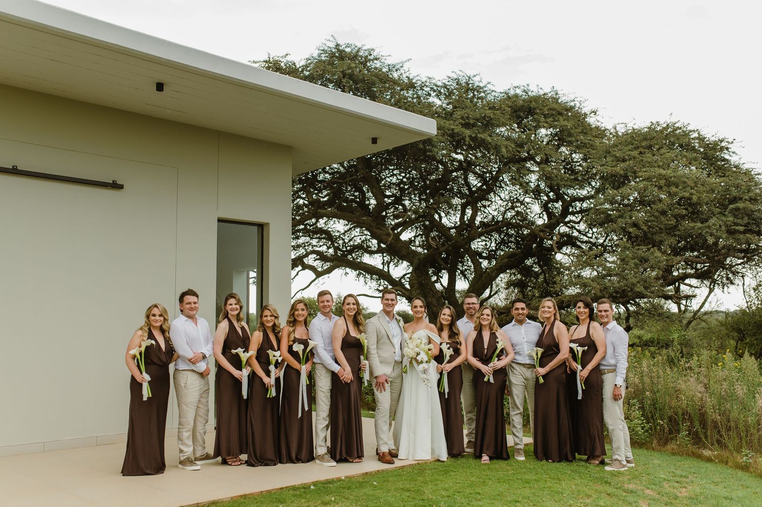 Wedding party poses together outdoors under a large tree beside a modern white building.