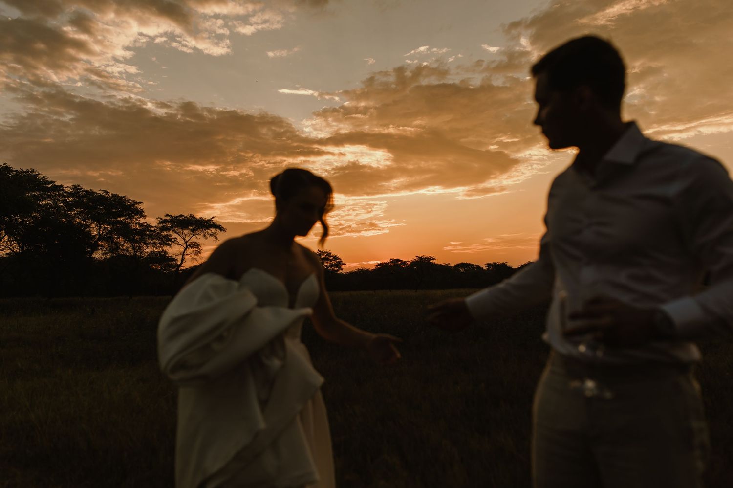 Silhouettes of a wedding couple embracing during a dramatic sunset in an open field.