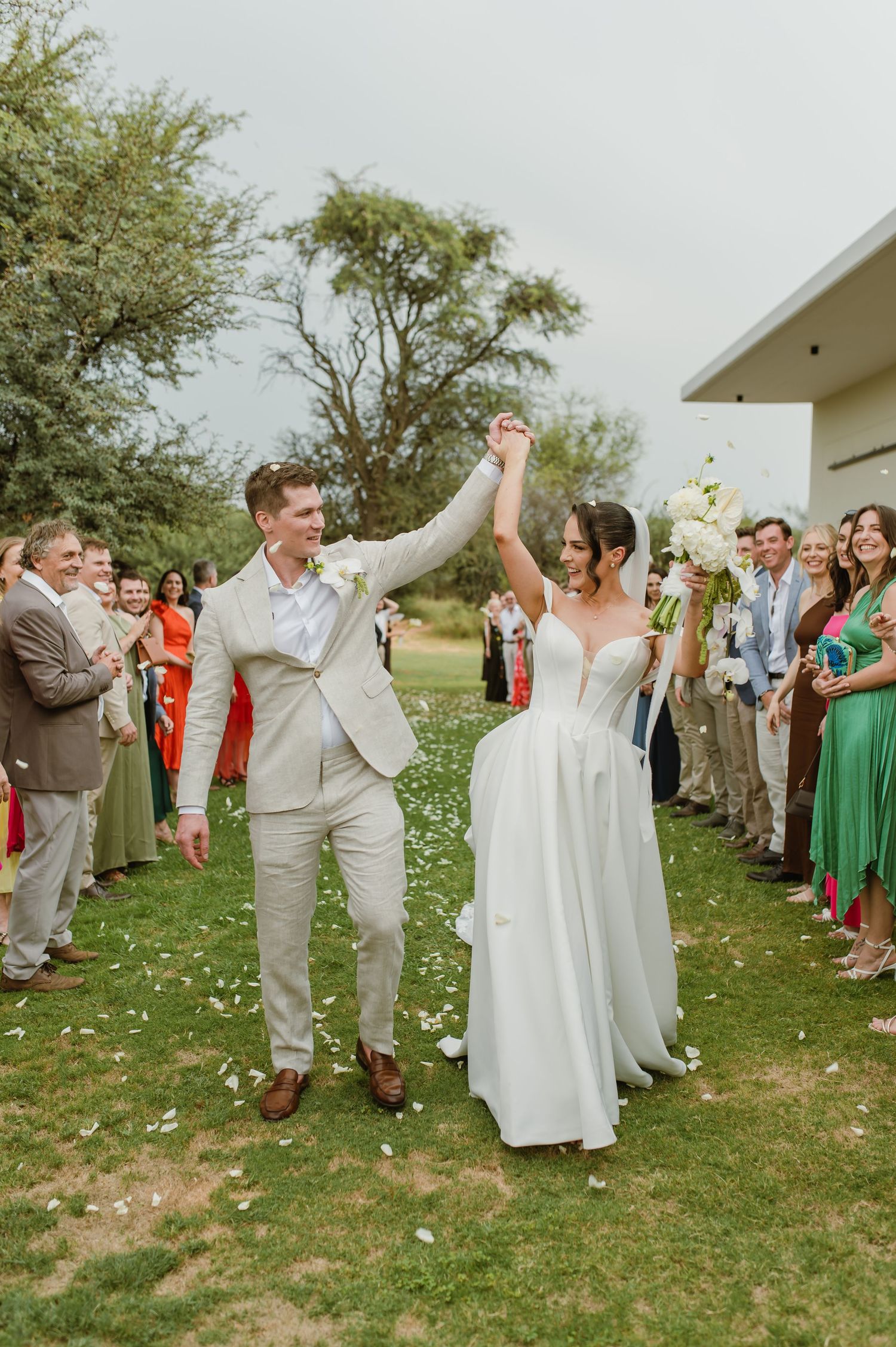 Newlyweds celebrate with raised hands on grassy lawn surrounded by applauding wedding guests.