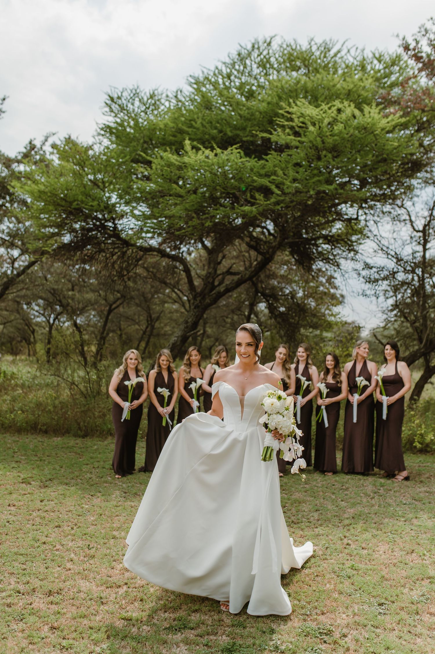 Wedding party in black dresses gathered outdoors under green trees for a group photo.