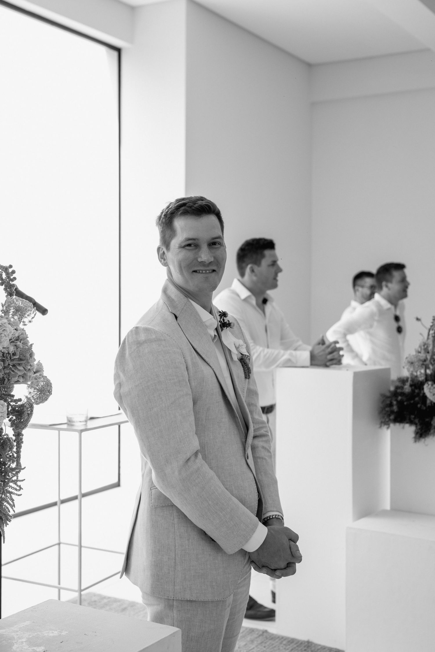 A groom in a light gray suit stands in a modern white venue during a wedding ceremony.