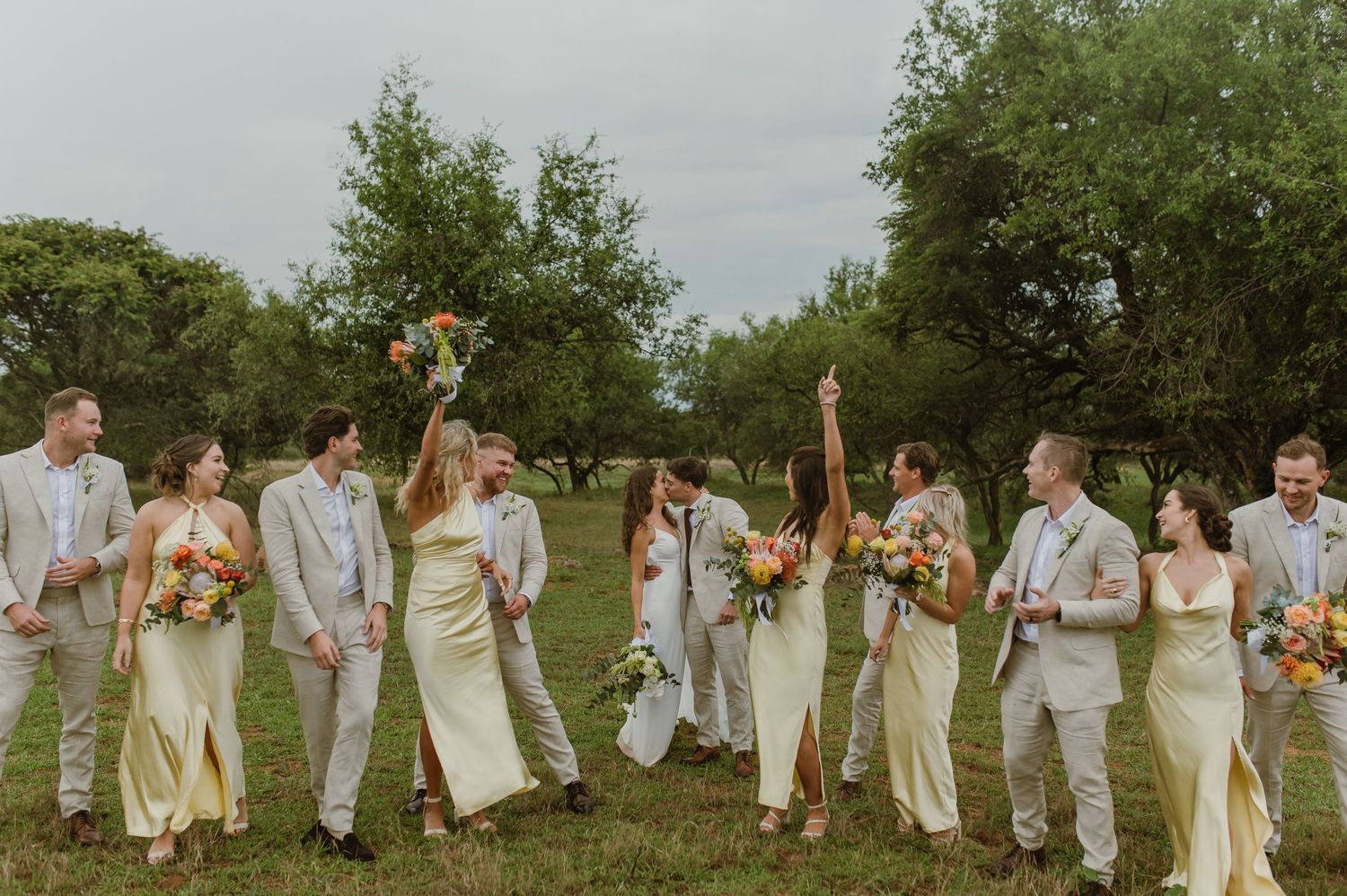 Wedding party members arranged in a long line outdoors wearing coordinated light-colored attire.