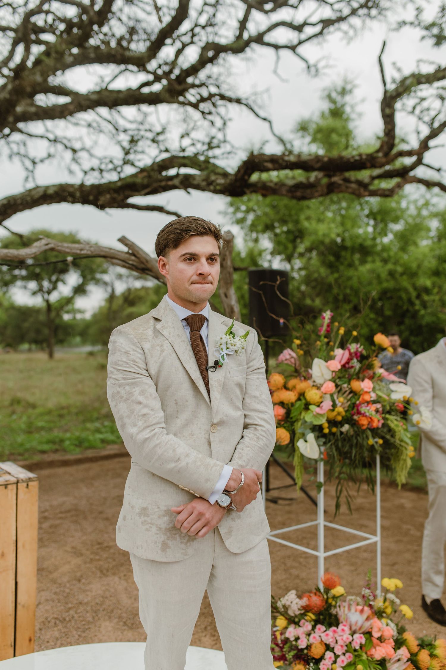 Groomsmen in light beige suits stand near floral arrangements under a large oak tree.