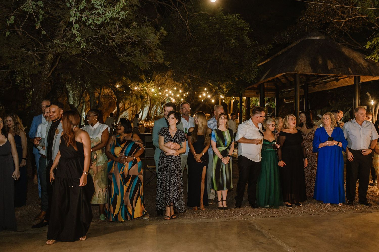 Large group of celebration attendees gathered for an outdoor evening event under trees.