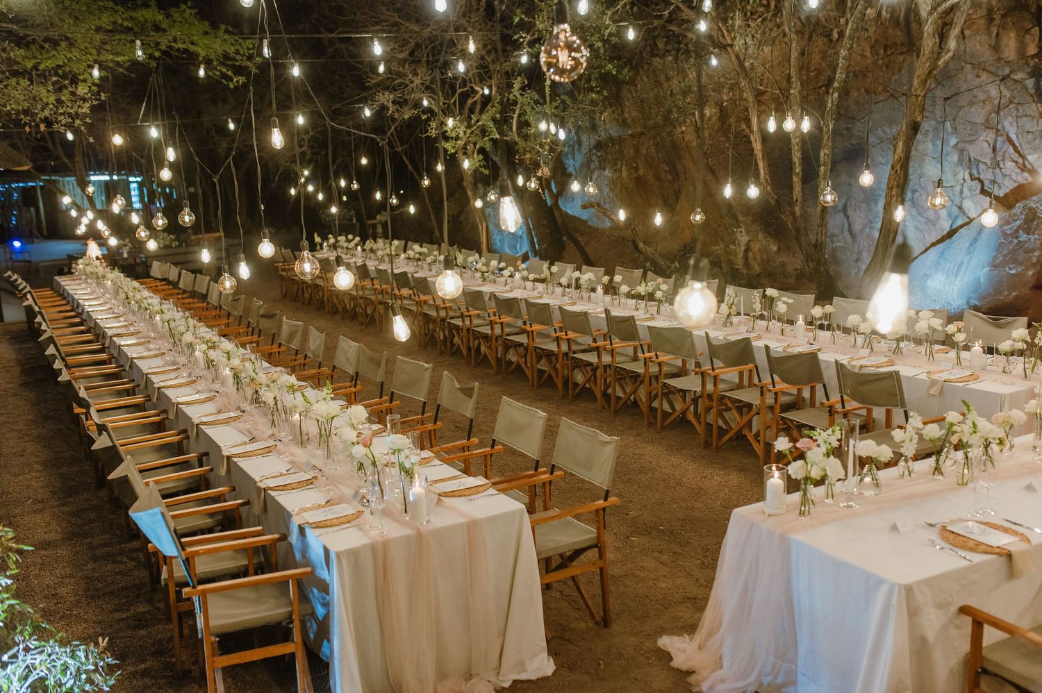 Evening outdoor wedding reception with wooden chairs and white tablecloths illuminated by string lights.