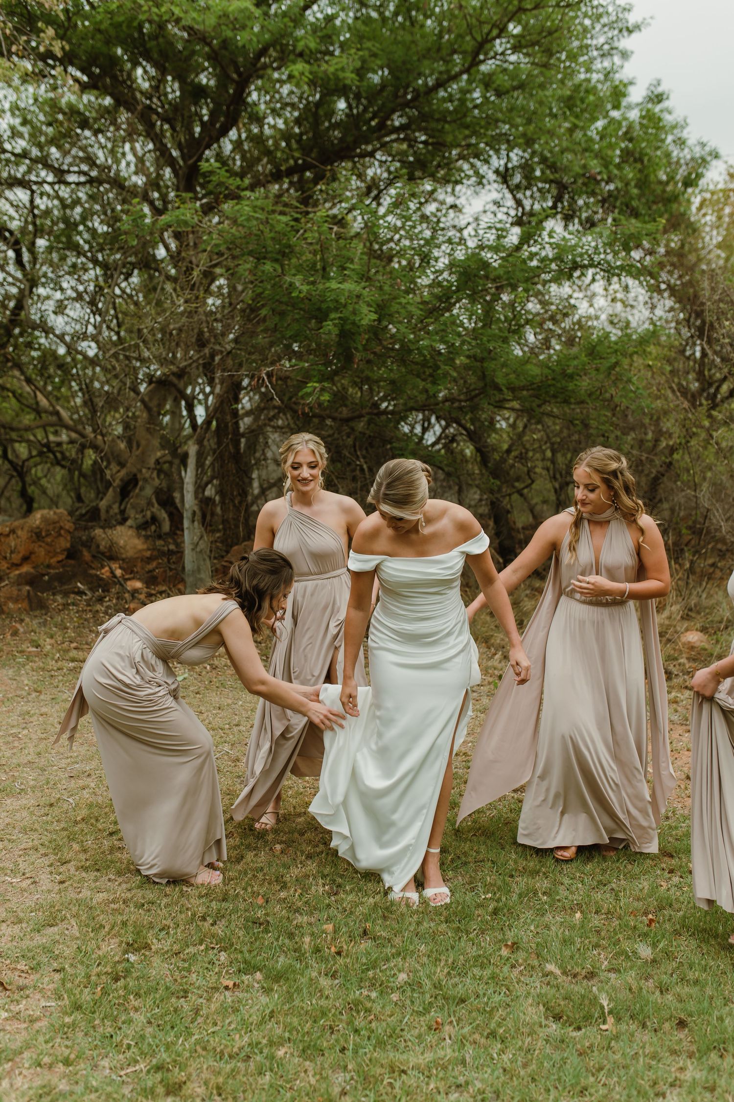 Bridesmaids in flowing beige dresses help adjust the bride's white gown on a grassy outdoor setting.