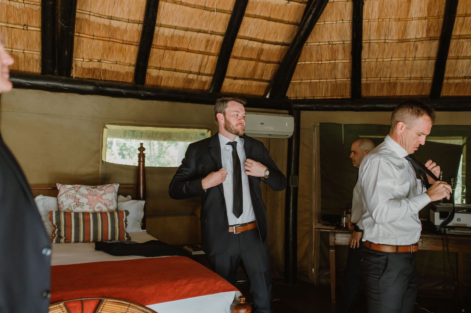 Wedding party members in dark suits gathered in a rustic venue with thatched roof.