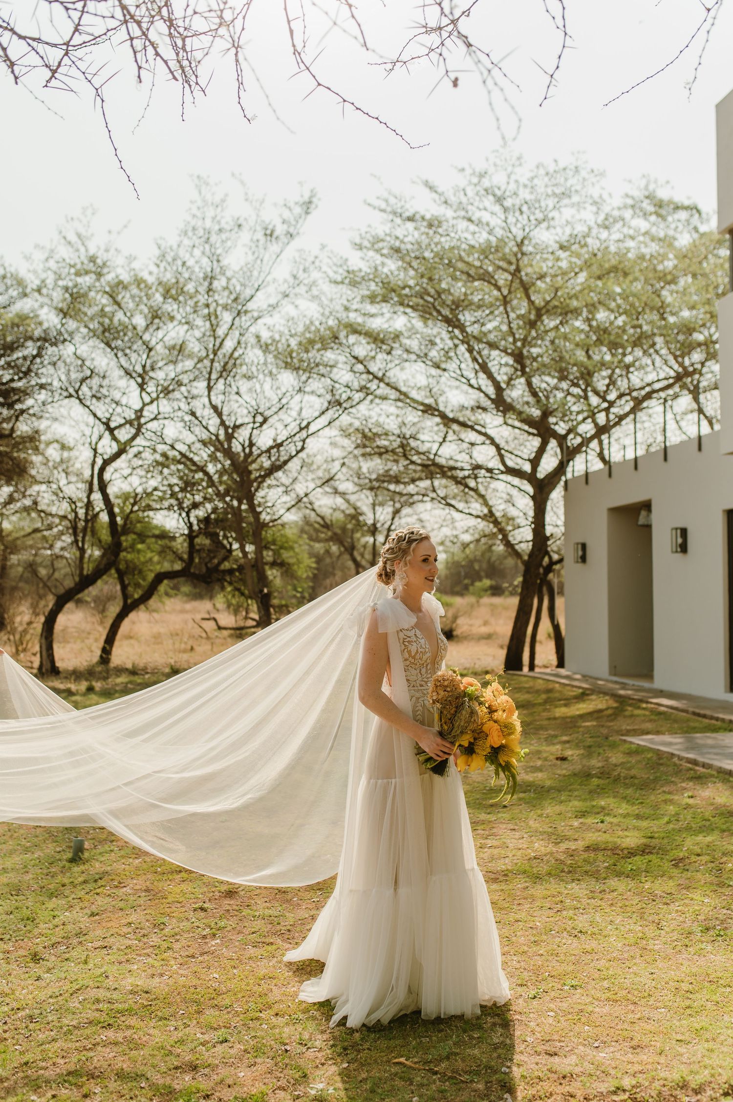 Bride with flowing veil holds sunflower bouquet against desert landscape and modern building.
