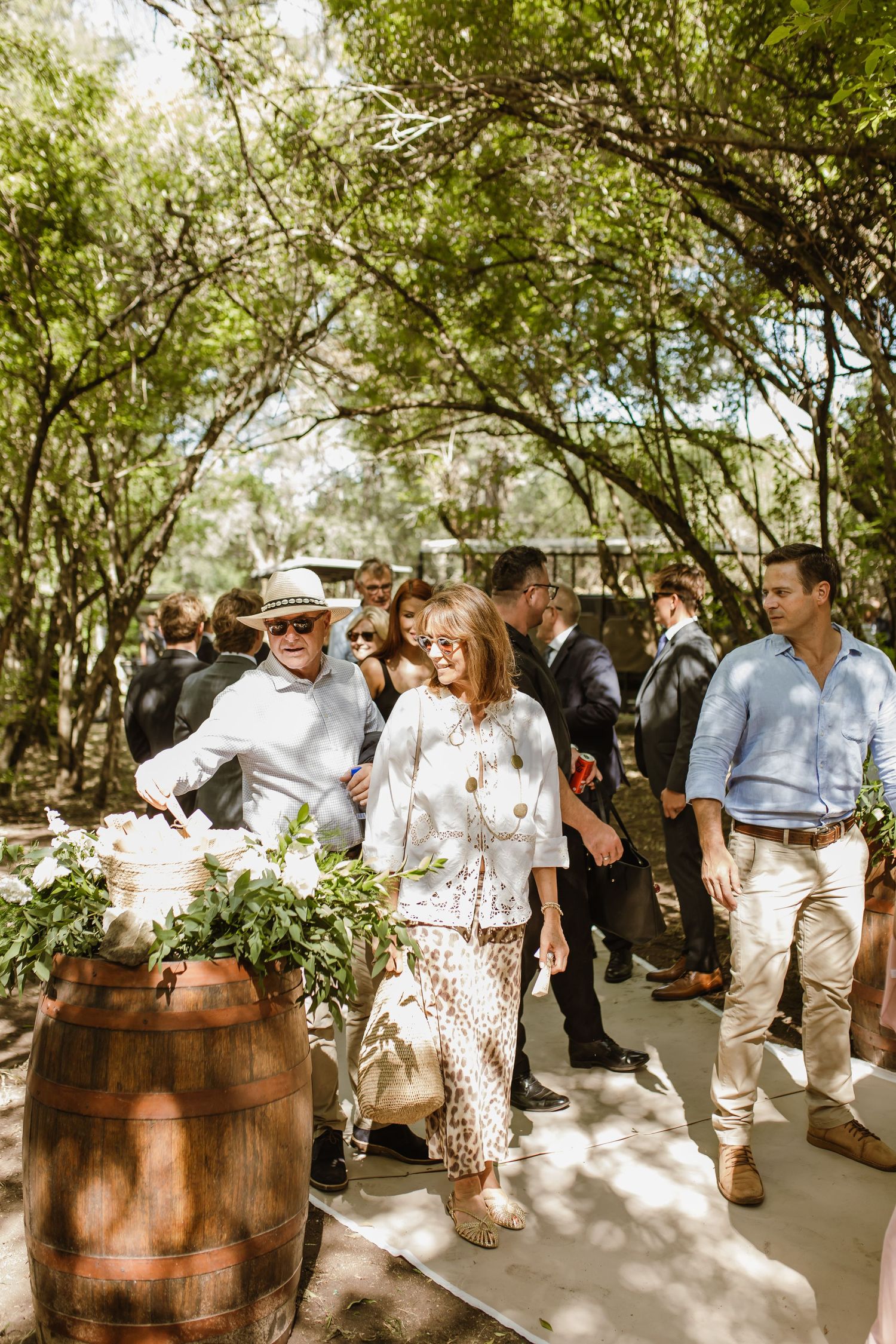 Wedding guests mingle near rustic wine barrels in a shaded outdoor garden setting.