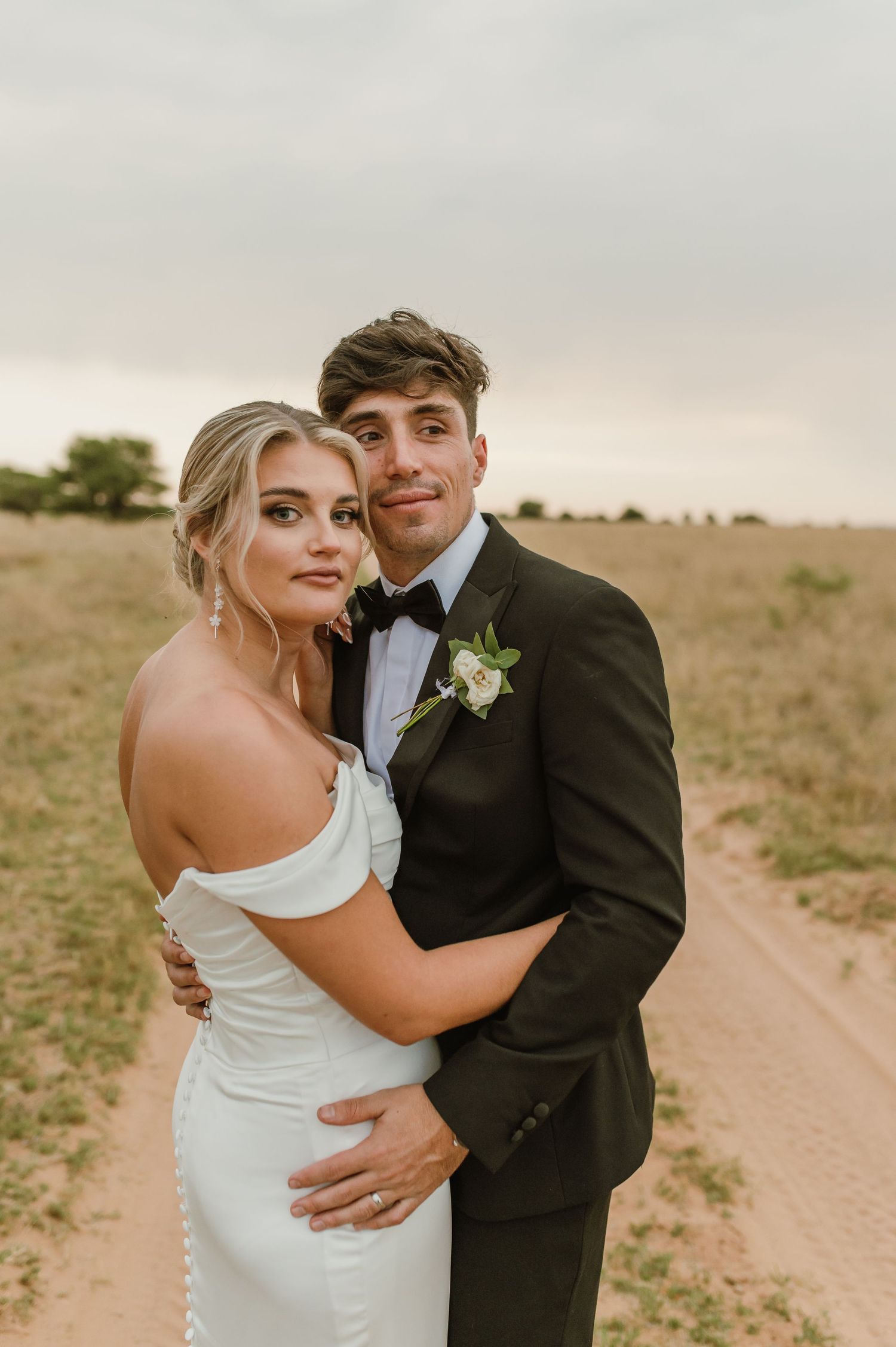 Newlywed couple embracing on a dirt path through an open field at sunset.