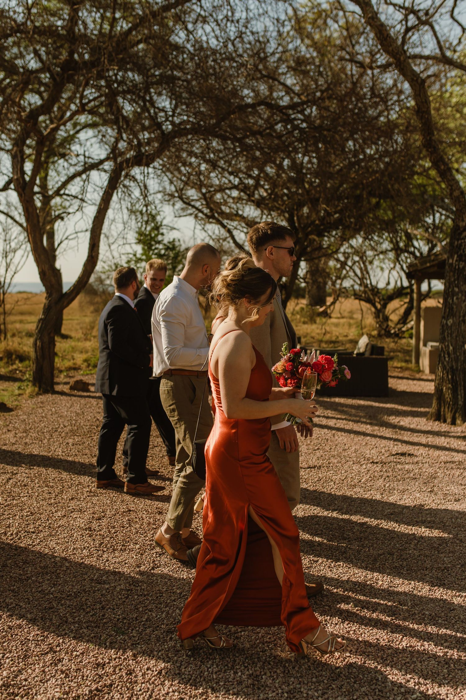 Wedding celebration on gravel path under shady trees during sunset.