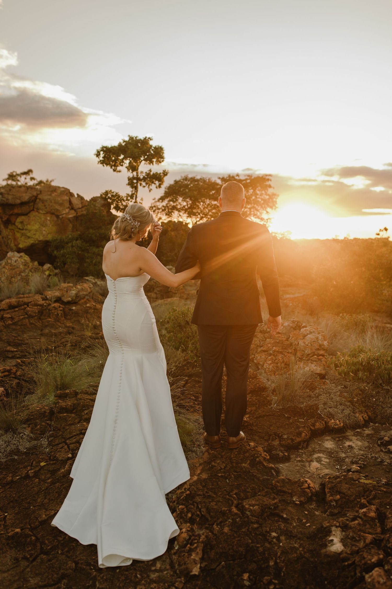 A couple in wedding attire walks along a rocky landscape at sunset with golden light illuminating the scene.