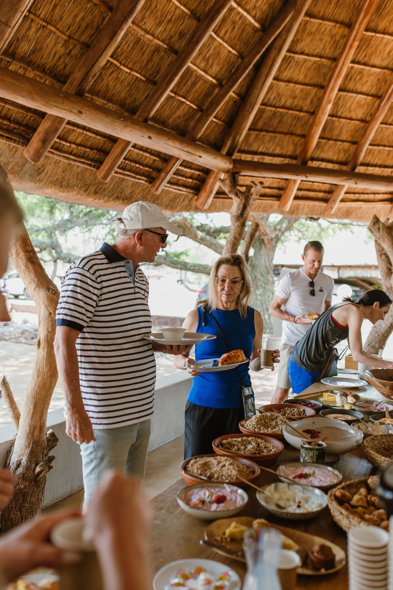 People serving themselves food from a buffet line under a thatched roof outdoor pavilion.