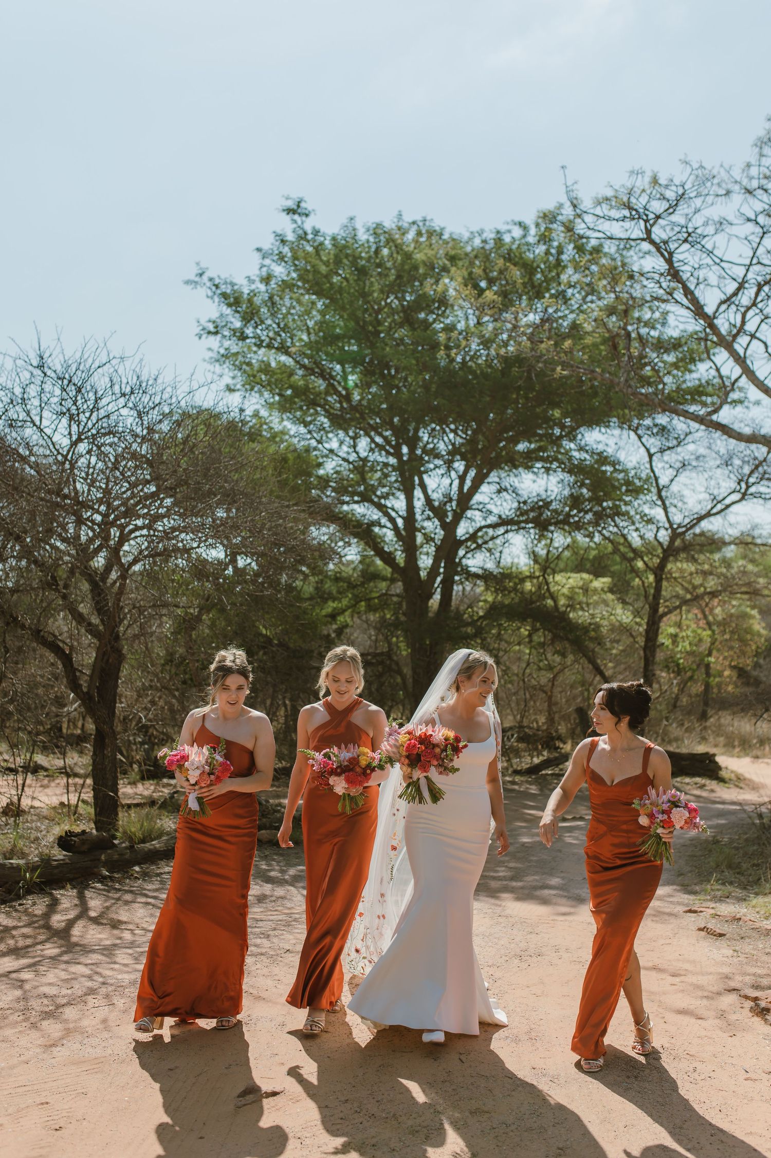 Bridesmaids in burnt orange dresses posing with bride in white gown outdoors.