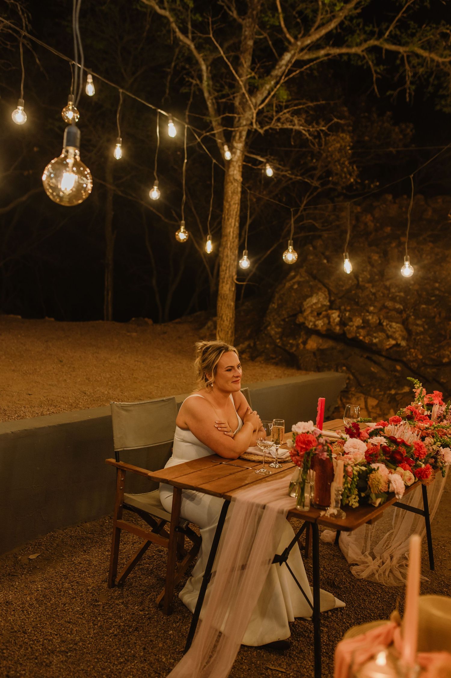Outdoor wedding reception table under string lights with floral centerpieces at dusk.