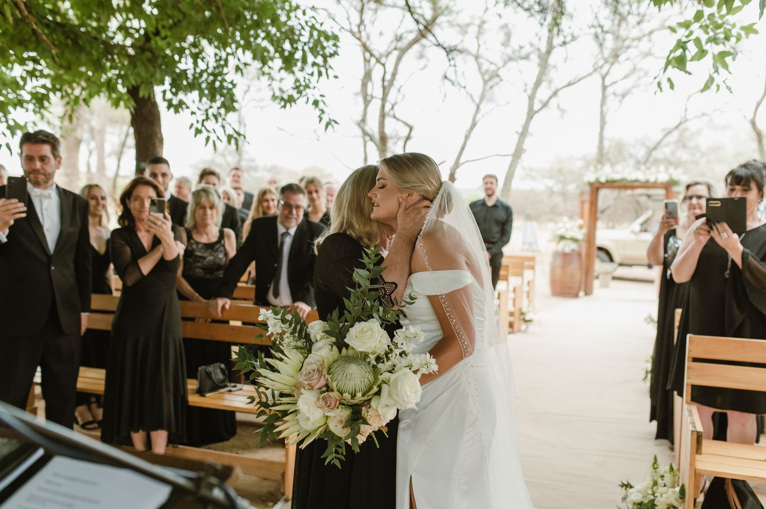 Elegant outdoor wedding ceremony with guests seated on wooden benches surrounded by trees.