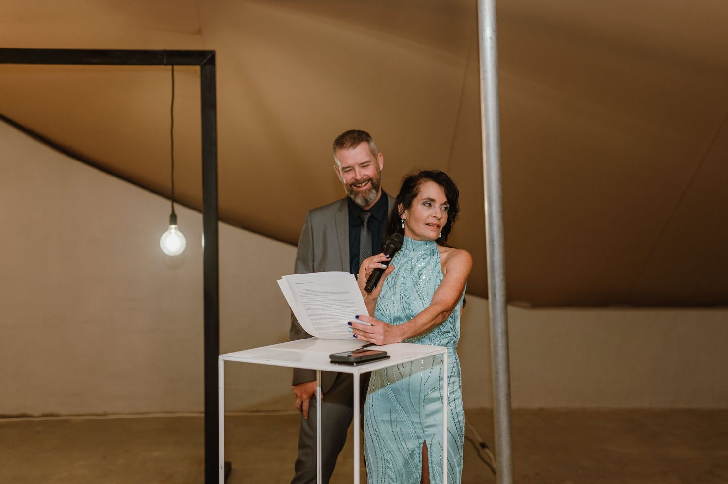 Wedding reception speeches delivered at head table under tent with ambient lighting.