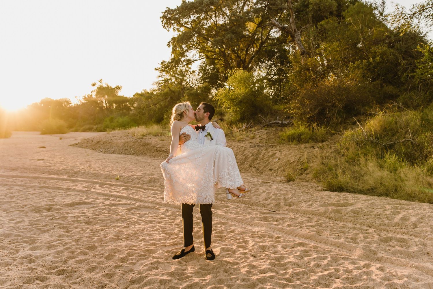 Groom carries bride in white dress across sandy beach at golden hour.