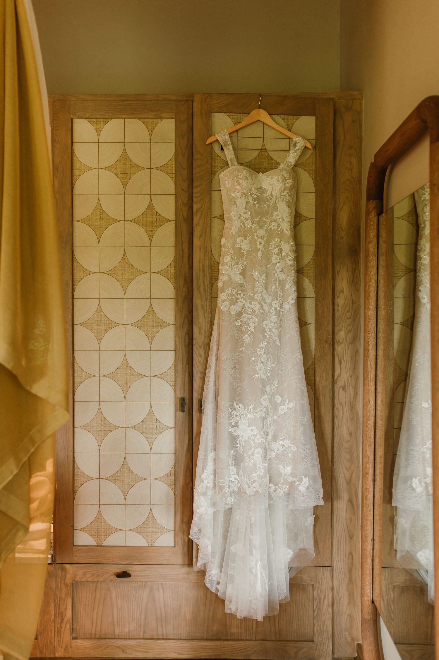 Elegant lace wedding dress hanging on a wooden wardrobe door in warm light.