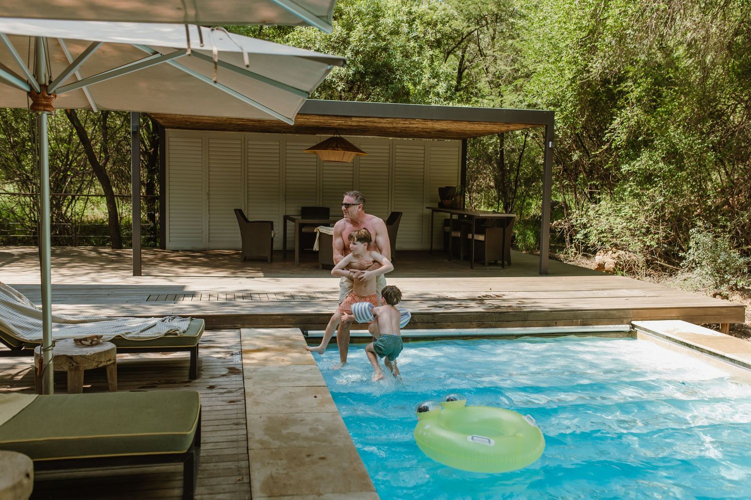 Family enjoying luxury safari lodge pool surrounded by lush trees with lounge area.