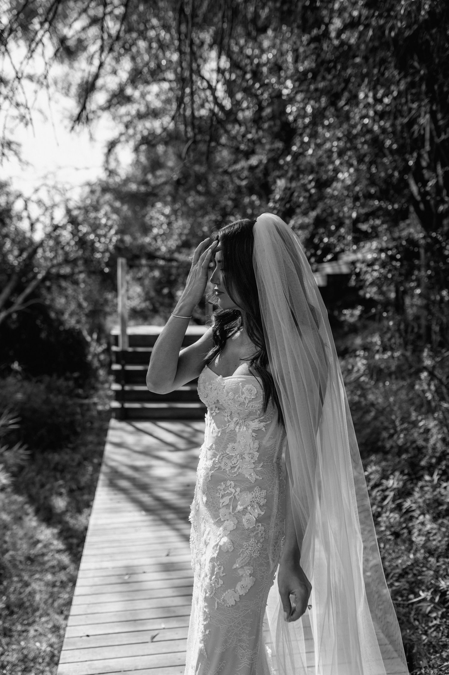 Black and white portrait of a bride in a lace gown and veil on a wooden bridge.