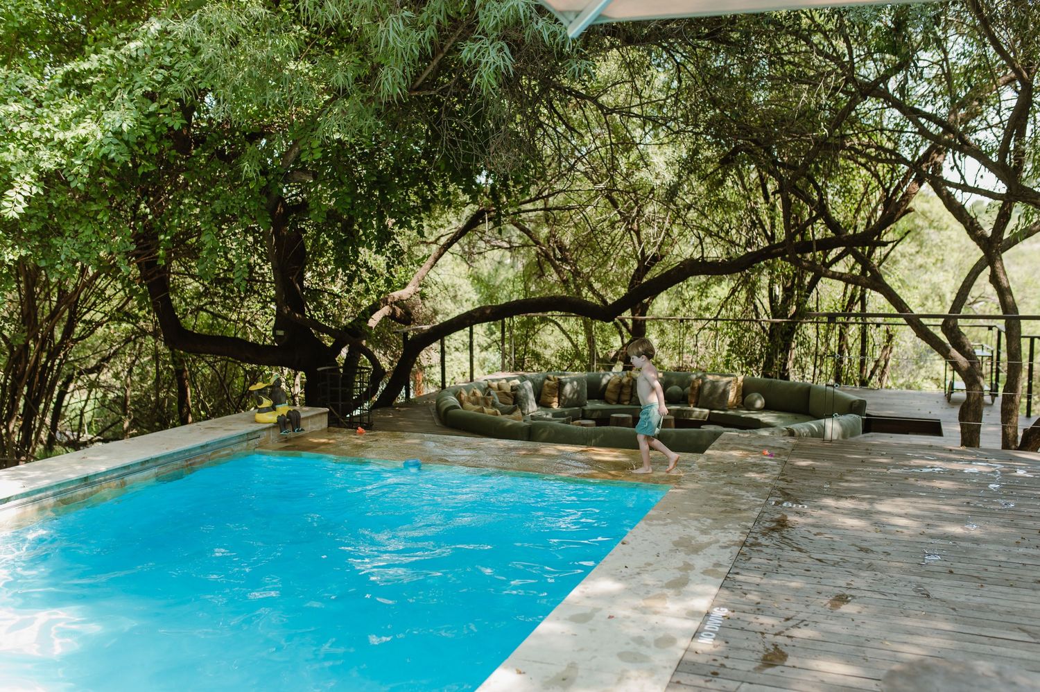 Person standing beside a blue outdoor pool surrounded by lush green trees on a wooden deck.