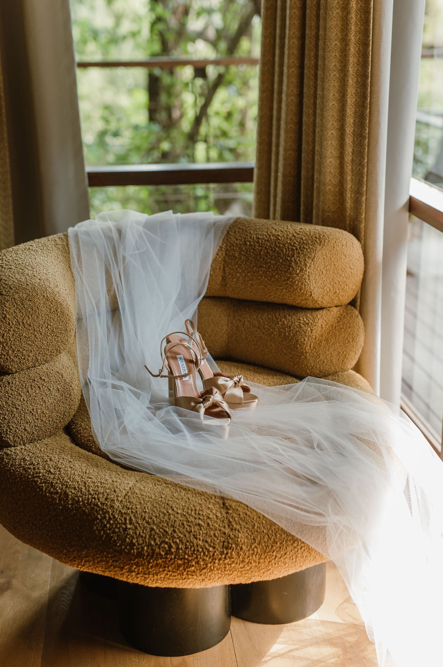 Bridal veil and strappy nude heels displayed on a modern boucle armchair near a sunlit window.