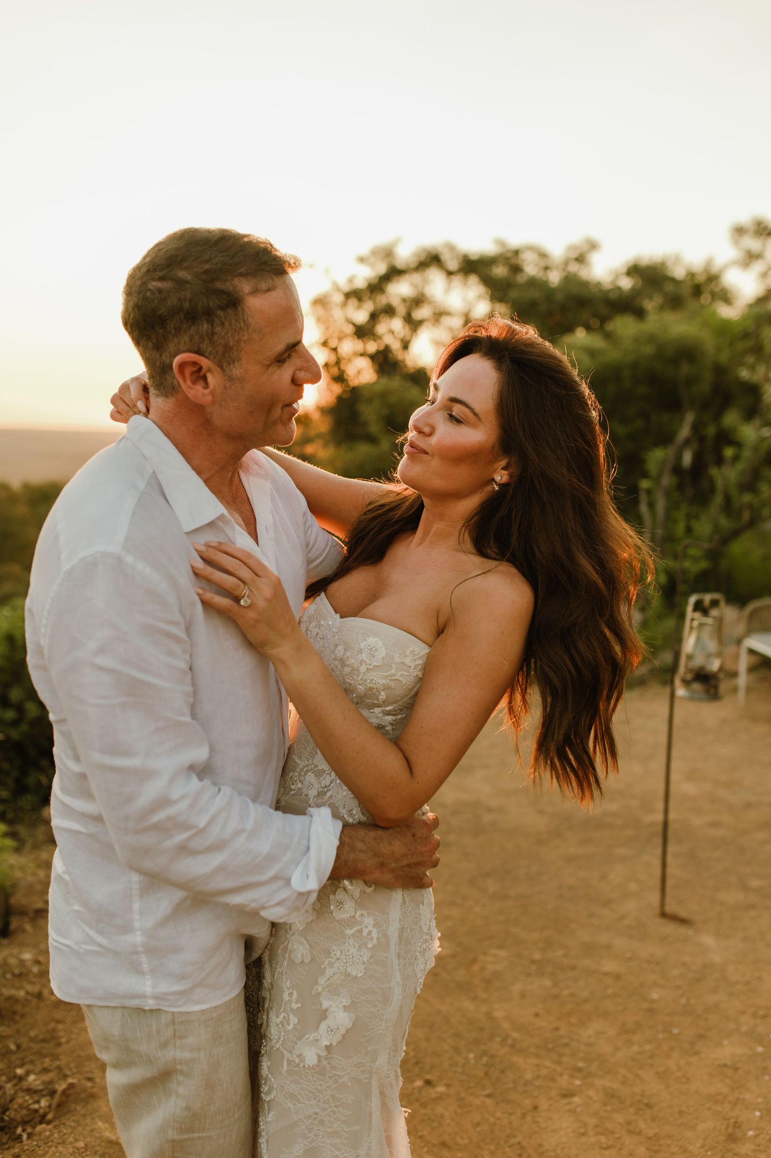 Bride and groom embrace at golden hour, she wears strapless lace gown, hair flowing in warm sunset light.