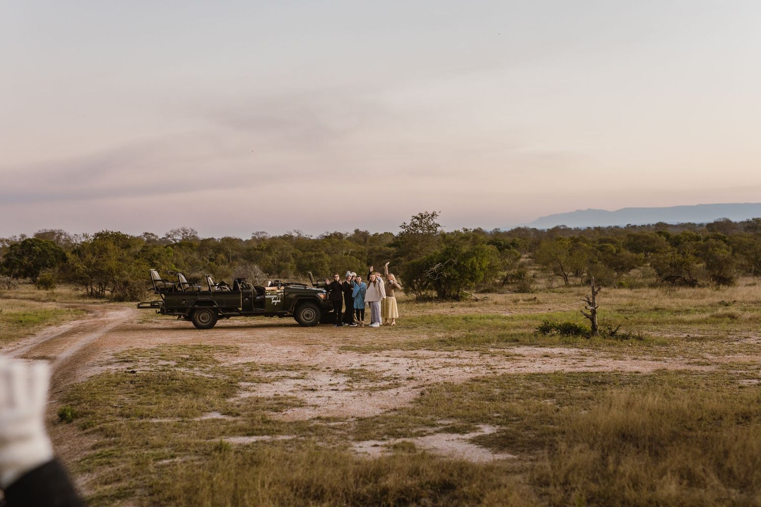 A group of people gather near a vehicle on a dusty path in a rural landscape at dusk.