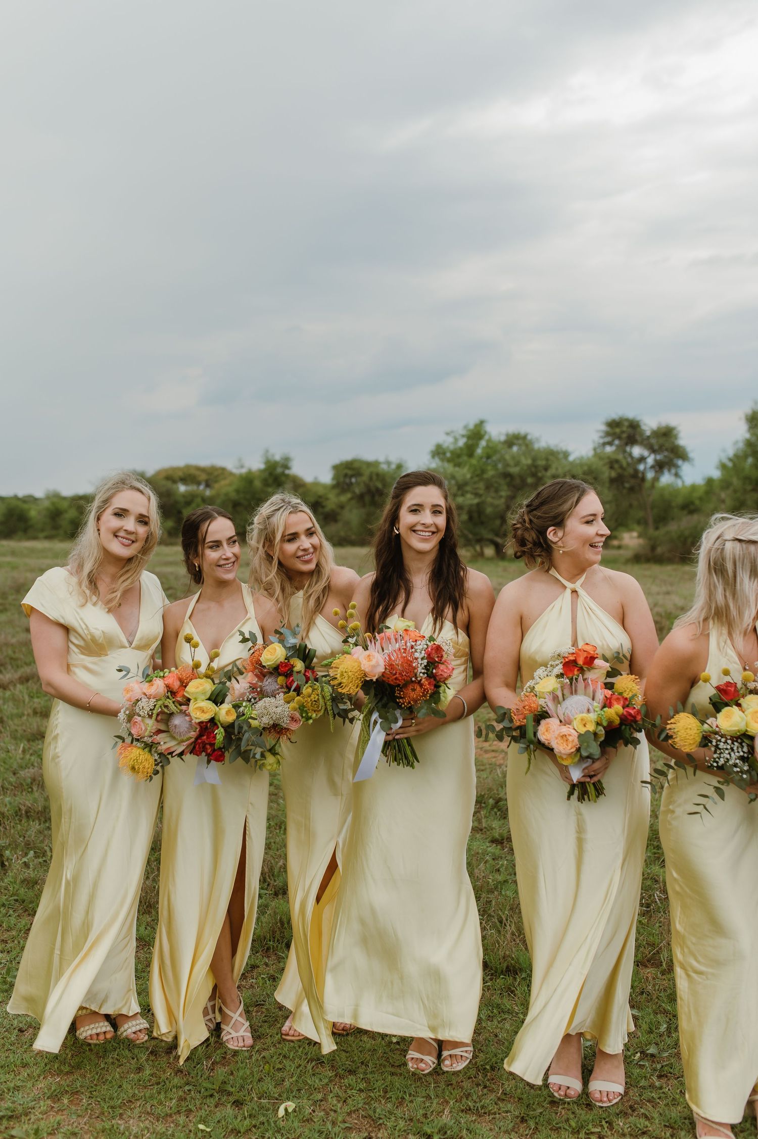 Bridesmaids in pale yellow dresses holding colorful bouquets pose together outdoors.