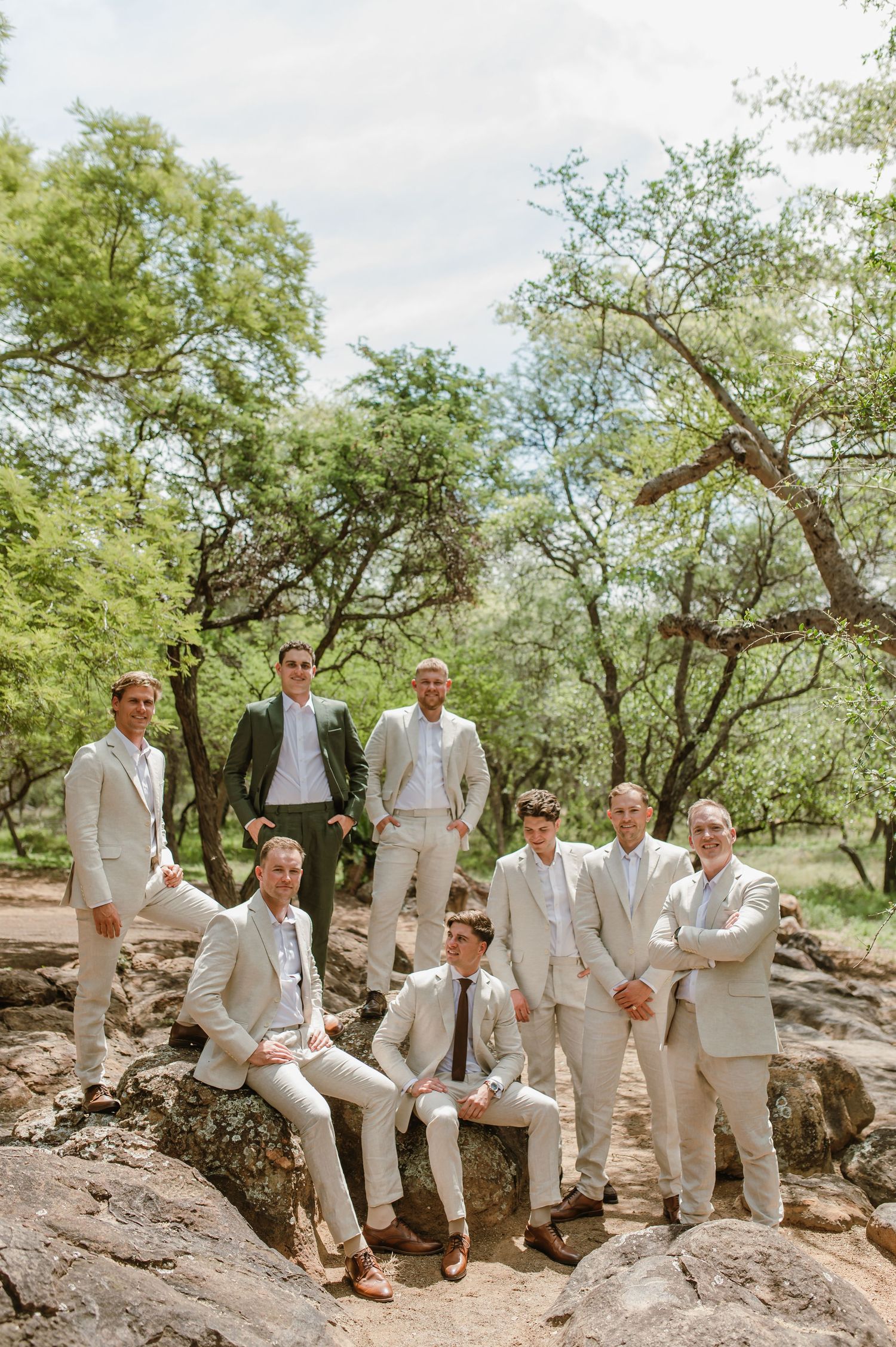 Groomsmen in beige suits pose casually on rocky terrain surrounded by trees.